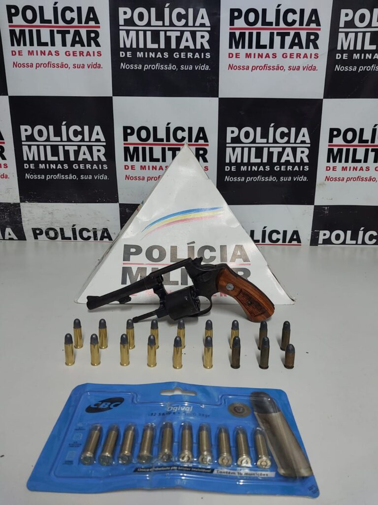 Violência Doméstica em Ponte Nova: Foto institucional da arma e munições apreendidas frente ao banner da polícia militar de minas gerais.