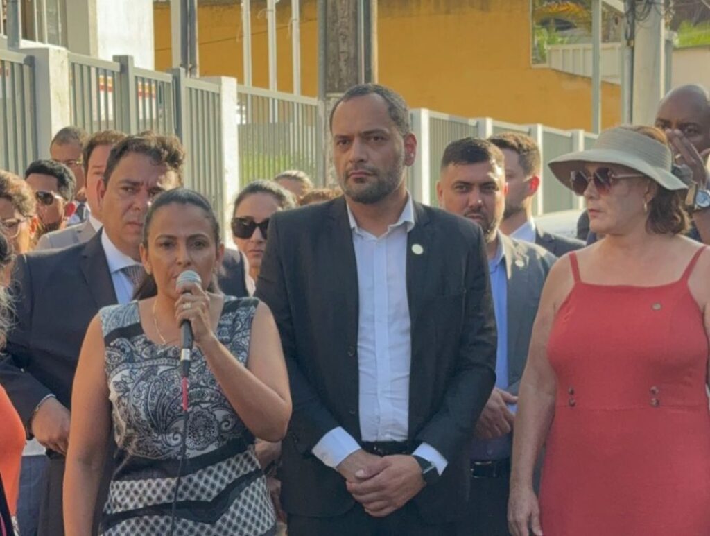 Ato de Desagravo:
Mulher baixa ao microfone, ao lado de homem mais alto de blaser preto, camisa social branca em posição de escuta e uma mulher com chapéu de proteção ao sol e vestido vermelho (vanja honorina).