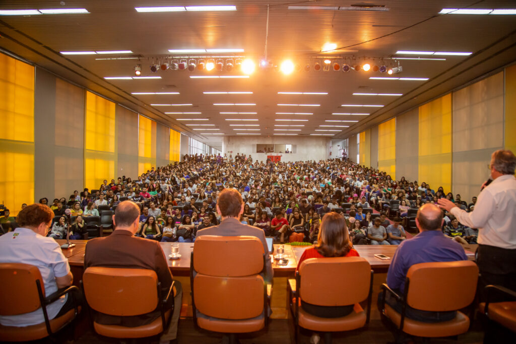 UFV Agendamento 2024 aberto
Foto institucional de um auditório grande de cadeiras vermelhas lotado. Vista de costas de mesa palestrante para o público.