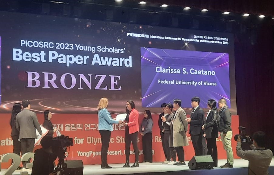 Pesquisa é premiada em conferência internacional

Vários homens coreanos de terno num palco grande, frente a um telão vibrante da premiação. Mulher de cabelos pretos lisos de blazer vermelho recebendo um certificado de papel de uma mulher loira, também de cabelos lisos, de blazer azul