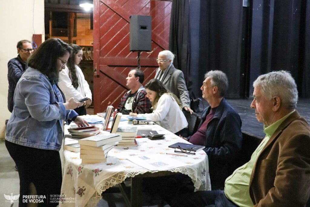 Festival Literário de Viçosa
Imagem ilustrativa da edição de 2023 de lançamento de livro no auditório da Estação Hervê Cordovil
Uma mesa repleta de livros dispostos de forma organizada e expositiva, quatro pessoas sentadas atrás da mesa e três pessoas em pé observando os livros.
