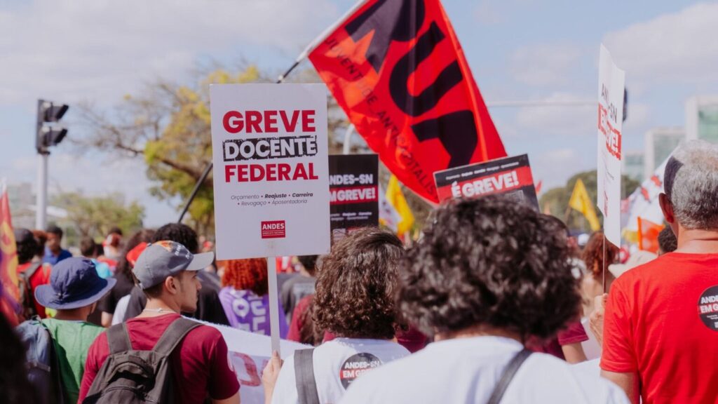 Uma multidão de pessoas participa de uma manifestação ao ar livre, a maioria vestida com camisetas vermelhas e carregando mochilas. Em destaque, há vários cartazes e bandeiras. O cartaz mais próximo da câmera tem os dizeres "GREVE DOCENTE FEDERAL" em letras vermelhas e pretas, seguido por "Orçamento • Reajuste • Carreira • Revogação • Aposentadoria" e o logo da ANDES (Sindicato Nacional dos Docentes das Instituições de Ensino Superior). Ao fundo, outras bandeiras e cartazes são visíveis, incluindo uma grande bandeira vermelha com letras pretas e o texto "ANDES-SN EM GREVE!". As pessoas estão reunidas em um espaço aberto, e ao fundo, é possível ver algumas árvores e edifícios em um dia claro e ensolarado.

Fim da greve dos professores