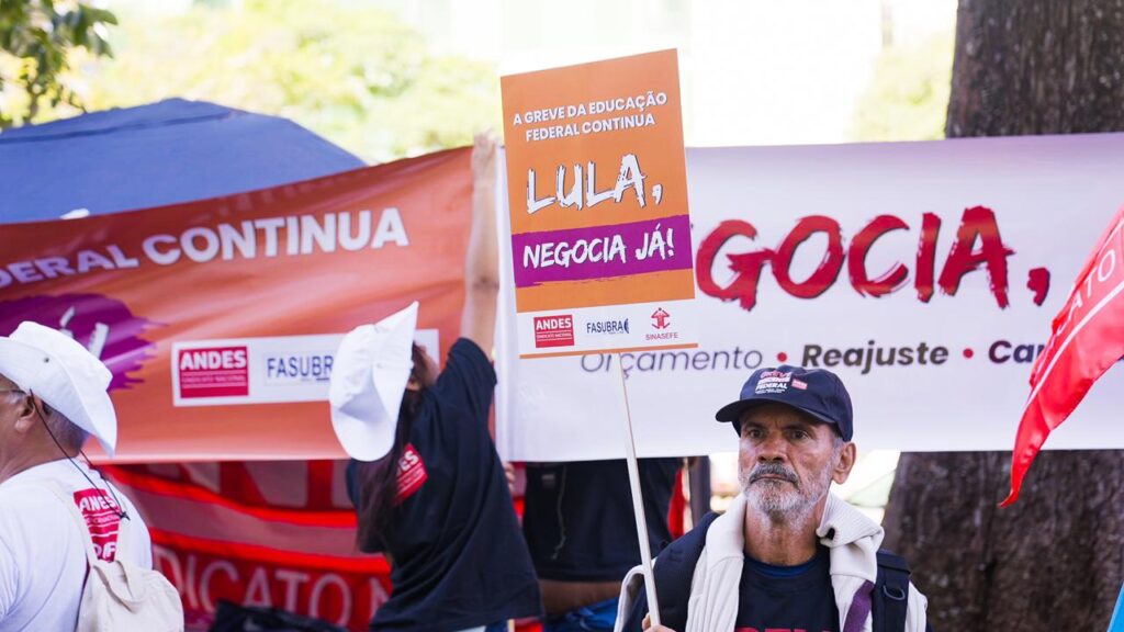 Governo rejeita contraproposta do ANDES mas docentes seguem pressionando em ocupações:
Imagem de um protesto de trabalhadores da educação federal. No centro, um homem com barba grisalha, usando boné preto e carregando uma placa laranja com as palavras "Lula, negocia já!" em letras brancas e roxas. A placa também inclui os logos do ANDES (Sindicato Nacional dos Docentes das Instituições de Ensino Superior), FASUBRA (Federação de Sindicatos de Trabalhadores Técnico-Administrativos em Instituições de Ensino Superior Públicas do Brasil) e SINASEFE (Sindicato Nacional dos Servidores Federais da Educação Básica, Profissional e Tecnológica). Ao fundo, outras pessoas seguram faixas vermelhas e brancas com frases que destacam a continuidade da greve e reivindicações como "orçamento", "reajuste" e "carreira". Algumas pessoas usam chapéus brancos, e há uma árvore e vegetação ao fundo, sugerindo que a manifestação ocorre ao ar livre.