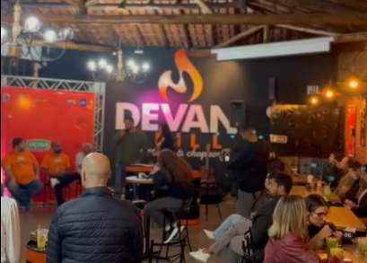 Festival Abrasel de Gastronomia e Cultura: Imagem de um espaço interno de um restaurante ou bar chamado "Devan Grill & Choperia". No centro da imagem, há um homem de pé, segurando um microfone e falando para o público, ele é o secretário de fazenda, presente no lançamento de um evento gastronômico. À esquerda, há um palco com fundo vermelho e algumas pessoas sentadas em cadeiras, observando o orador. Ao redor, várias mesas ocupadas por pessoas sentadas, conversando e prestigiando o evento. A decoração inclui luminárias penduradas e uma estrutura de madeira exposta no teto. A atmosfera é animada e acolhedora, com luzes quentes iluminando o local.