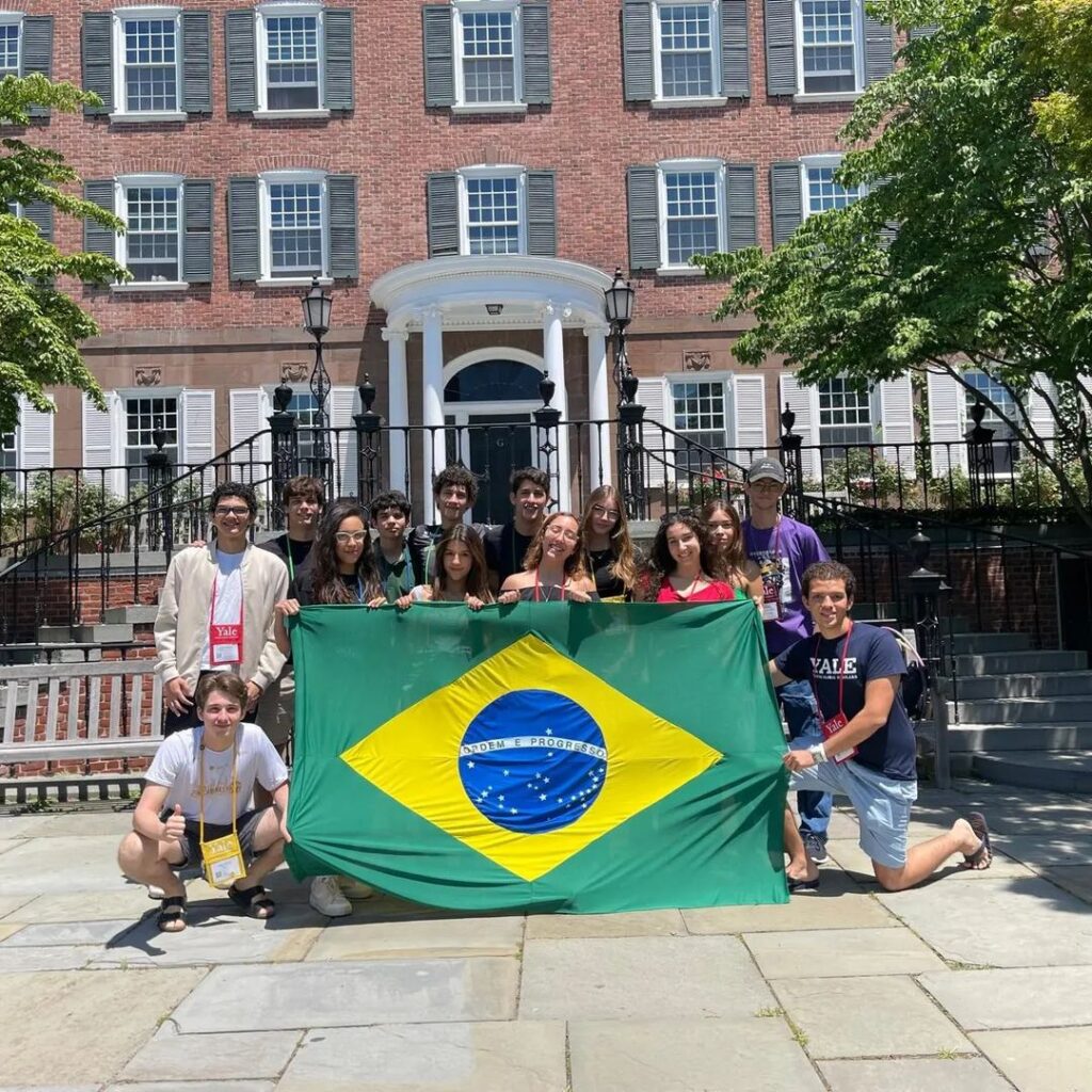 Aluna do Coluni representa Brasil internacionalmente;

A imagem mostra um grupo de jovens em frente a um edifício de tijolos vermelhos com várias janelas, todas com venezianas cinza, e uma entrada central com uma varanda e colunas brancas. Os jovens estão posando juntos, segurando uma grande bandeira do Brasil, que é verde com um losango amarelo e um círculo azul estrelado no centro com a inscrição "Ordem e Progresso" em uma faixa branca.

Na parte inferior esquerda da imagem, um jovem está agachado, vestindo uma camiseta branca e segurando um crachá amarelo com o polegar para cima. Outro jovem, à direita, está ajoelhado, usando uma camiseta azul escura com o logotipo da Yale e segurando o lado direito da bandeira.

O grupo está sorrindo e parece estar participando de um evento acadêmico ou cultural. O dia está ensolarado e há árvores com folhagem verde ao redor do edifício.